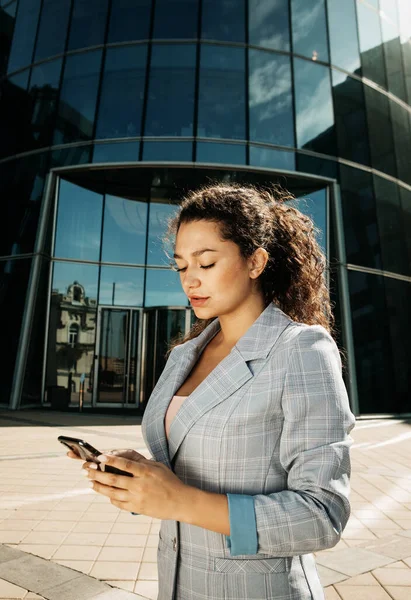 Joven mujer de negocios utiliza un teléfono móvil frente a un moderno centro de negocios —  Fotos de Stock