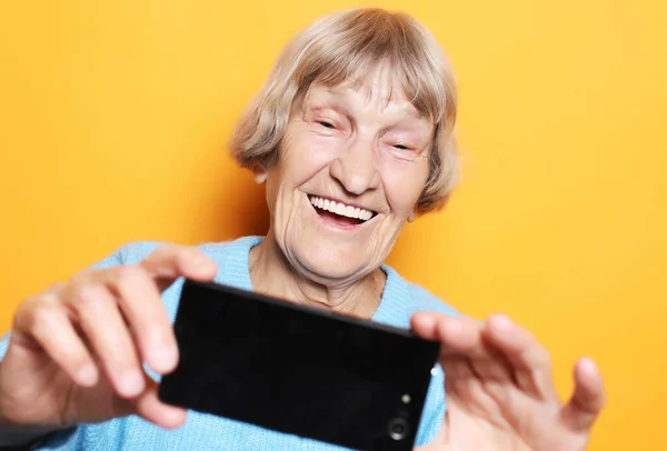 Lifestyle, tehnology and people concept: vovó sorri e toma uma selfie sobre fundo amarelo — Fotografia de Stock