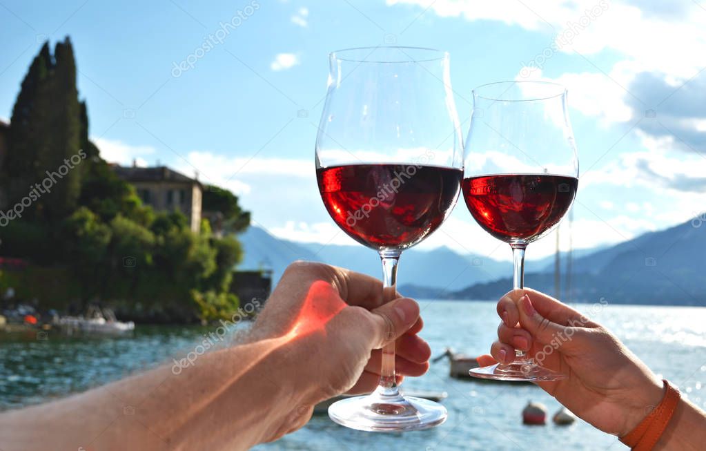 Hands holding two wineglasses. Varenna town at the lake Como, Italy
