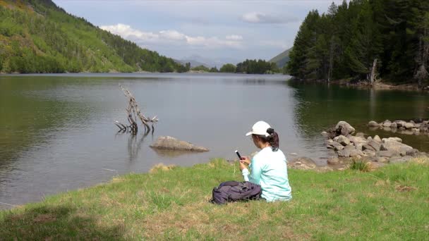 山の湖の隣に座って 携帯電話で写真を撮る若い観光少女 — ストック動画