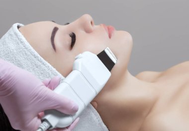 Doktor-kozmetik uzmanı cihazı güzellik salonundaki genç ve güzel bir kadının yüz derisinin ultrasonla temizlenmesi için bir prosedür haline getiriyor. Kozmetoloji ve profesyonel cilt bakımı.