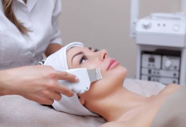 Doktor-kozmetik uzmanı cihazı güzellik salonundaki genç ve güzel bir kadının yüz derisinin ultrasonla temizlenmesi için bir prosedür haline getiriyor. Kozmetoloji ve profesyonel cilt bakımı.