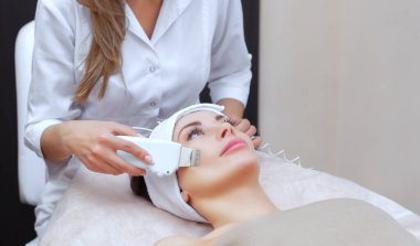 Doktor-kozmetik uzmanı cihazı güzellik salonundaki genç ve güzel bir kadının yüz derisinin ultrasonla temizlenmesi için bir prosedür haline getiriyor. Kozmetoloji ve profesyonel cilt bakımı.