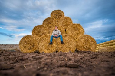 Bir adam mavi gökyüzüne karşı sarı saman rulolarında oturur.