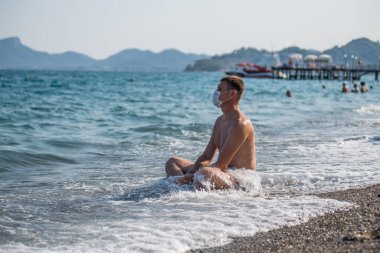 19 numaralı grip salgını sırasında plajda maske takan bir adam tek başına oturuyor.