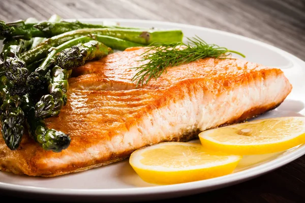 Roasted salmon fillet with baked asparagus and sliced lemon on white plate on wooden table