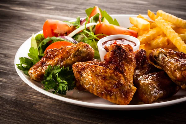 Roasted chicken wings with wavy french fries and fresh vegetables on white plate on wooden table