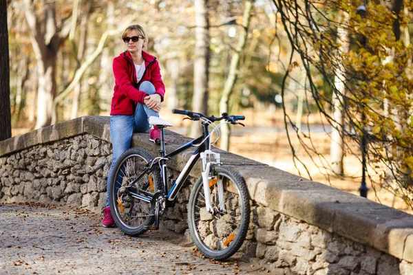 在秋天的公园坐在石头栅栏上的成年妇女 在前景上骑自行车 — 图库照片