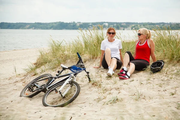 两名戴着太阳镜的中年妇女在自行车附近的海边休息 — 图库照片