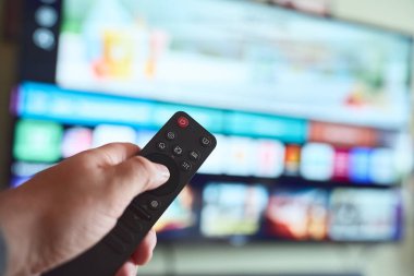 Geniş bir TV ekranında çeşitli yayın seçeneklerini gösteren programı seçerken uzaktan kumandayı tutan bir el.