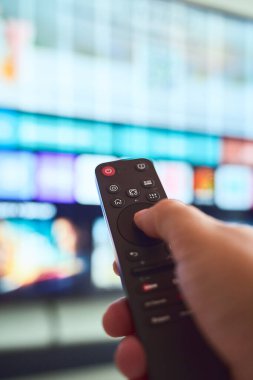 Bir el, televizyon ekranında çeşitli renkli kanal ve görüntüleri gösteren seçenekleri aktif olarak seçen bir uzaktan kumanda tutar.