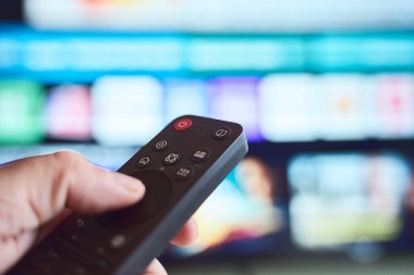 Akşamları elinde uzaktan kumandayla farklı televizyon kanallarına girip eğlence seçeneklerini keşfeden bir kişi....