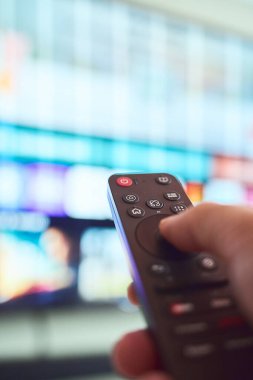 Bir el, rahat bir oturma odasında renkli içeriklerle dolu büyük bir ekranda kanalları tararken uzaktan kumandayı tutuyor..