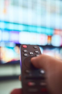 Bir insan, oturma odasındaki parlak bir televizyon ekranında uzaktan kumandayı kullanarak kanalları gezer..