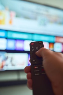 Modern bir oturma odasında renkli bir televizyon ekranı izlerken uzaktan kumandalı, kanal değiştiren bir insan..