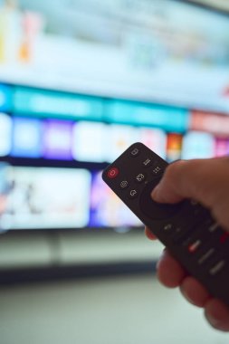 Bir el, rahat bir oturma odasında renkli uygulama simgeleriyle dolu akıllı bir TV arayüzünü kullanırken uzaktan kumandayı tutar.