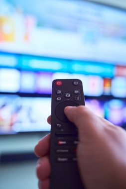 Bir el, oturma odasında çeşitli yayın seçeneklerini gösteren akıllı bir televizyona odaklanırken uzaktan kumandayı tutuyor..