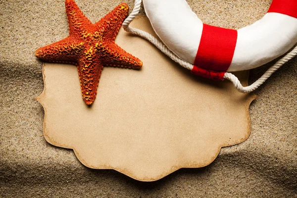Orange starfish, red and white lifebuoy and blank paper label on beach sand