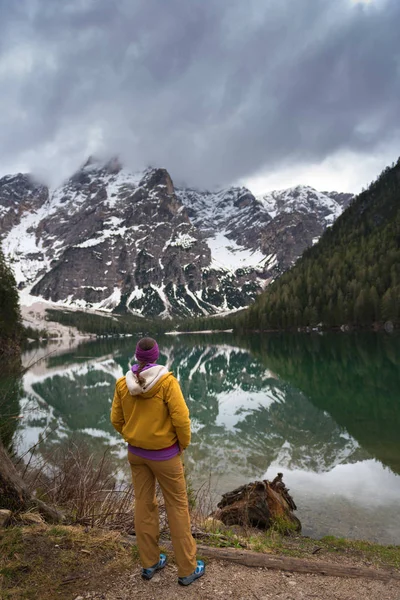 A brunette woman hiker, wearing outdoor gear is standing looking out at the view on Lake Braies. Golden and green trees reflect the autumn.