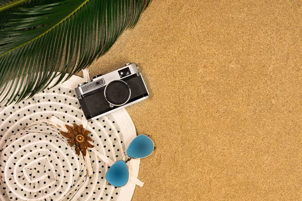 sunglasses, hat, vintage camera on sand background, top view