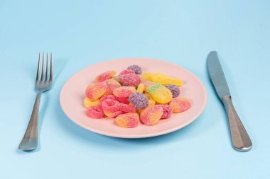 Mavi arka plan üzerinde izole sakızlı şekerler ile çatal, bıçak ve pembe plaka
