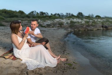 Plajda hamile çift, kum üzerinde oturan ve kavun yeme. 