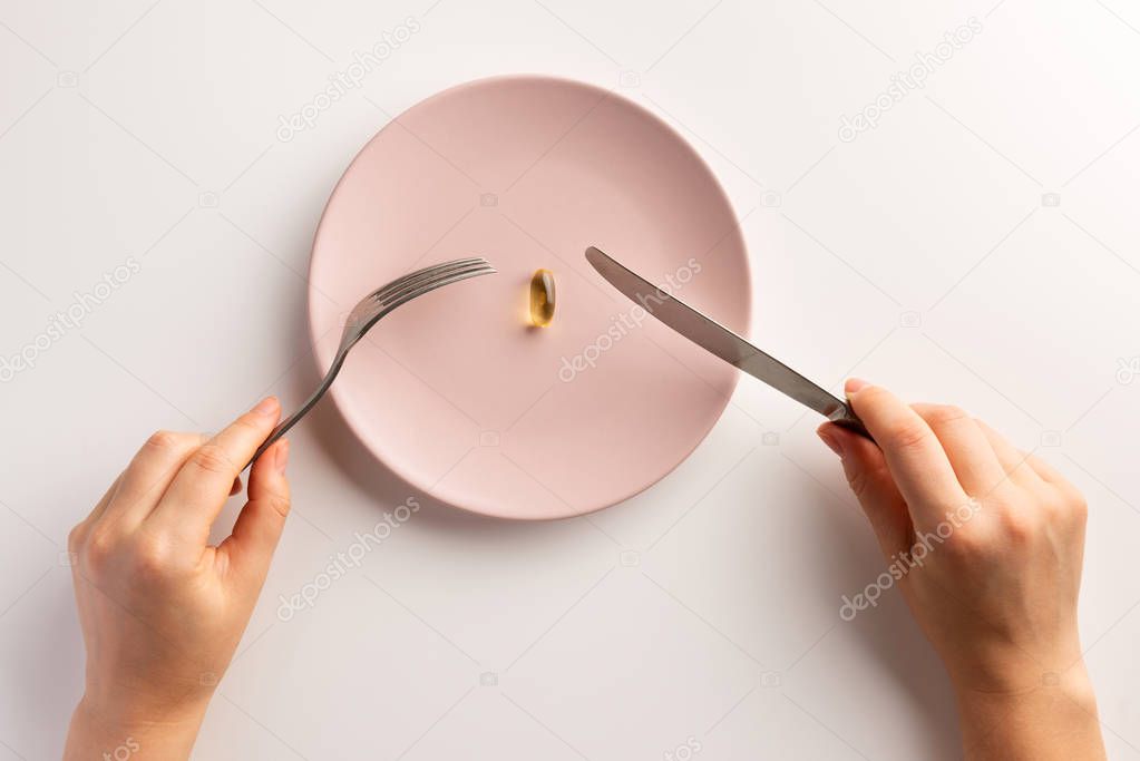 Female hands holding fork and knife. Plate with fish oil capsule. Flat lay.