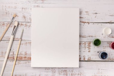 Çizimler ve boyalar için boş tuvali olan renkli yaratıcı masa, beyaz ahşap masaya dizilmiş boya fırçaları.