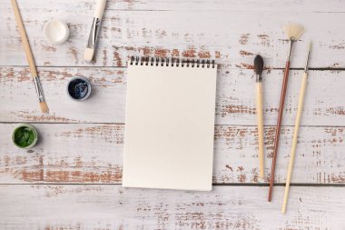 Sanatçının işyeri. Beyaz ahşap masadaki fırçalı resimler ve boş eskiz defterleri, üst görünüm, düz alan, nesneler