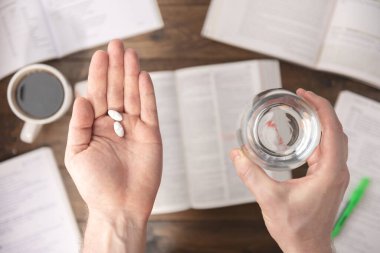 İş stresi konsepti. Çalışma masasının üzerinde ilaç, kitap, kalem, kağıt ve bir fincan kahve taşıyan eller. 