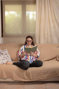 Evdeki genç kadın pencerenin önündeki koltukta oturuyor oturma odasında kitap okuyor. 