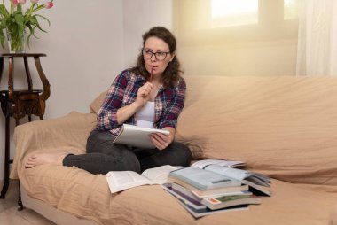 Koyu kıvırcık saçlı genç ve çekici bir kadın kanepeye oturmuş elinde defterler ve kitaplarla modern bir evde ders çalışıyor.