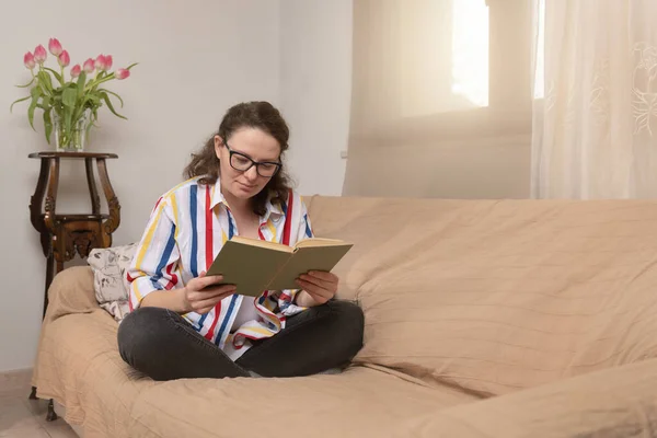 Calm satisfied Caucasian woman relaxing on comfortable couch alone, holding paper book, spending weekend, free time at home with literature