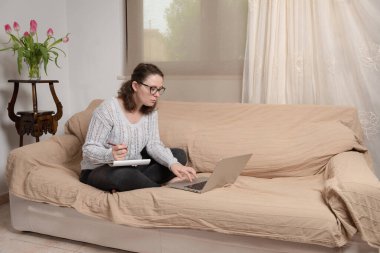 Koyu kıvırcık saçlı genç ve çekici bir kadın kanepede dizüstü bilgisayar ve kitaplarla oturuyor, modern bir evde ders çalışıyor. Evde çalışıyorum.