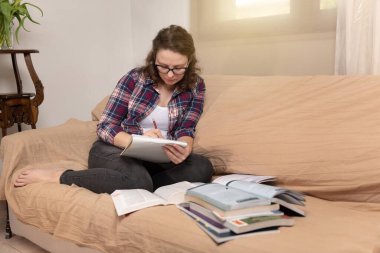 Koyu kıvırcık saçlı genç ve çekici bir kadın kanepeye oturmuş elinde defterler ve kitaplarla modern bir evde ders çalışıyor.