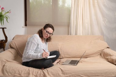 Oturma odasındaki kanepede oturan ve evde uzaktan çalışan genç bir bayan. Dizüstü bilgisayarı ve kitaplarıyla çalışıyor.