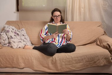 Evdeki genç kadın pencerenin önündeki koltukta oturuyor oturma odasında kitap okuyor. 