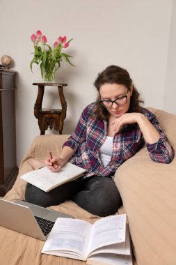 Kanepede oturan genç bir kız öğrenci, çalışırken kitap ve dizüstü bilgisayar kullanıyor.