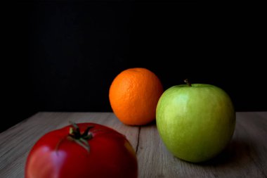 Olgun domates, yeşil elma ve portakal, siyah arka planda, tahta bir tahtanın üzerinde duruyor.