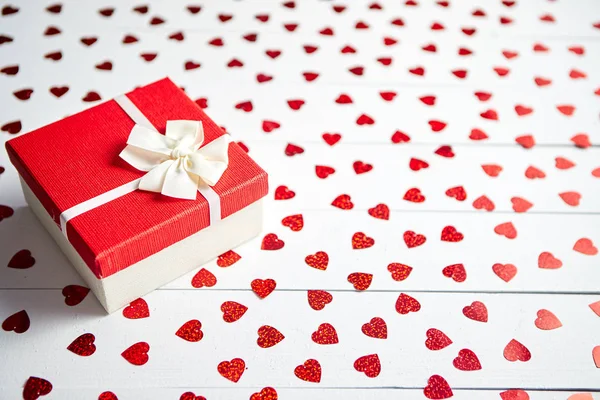 Cadeau en boîte placé sur des paillettes rouges en forme de coeur sur une table en bois blanc — Photo
