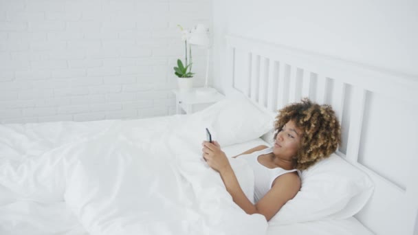 Mujer sonriente relajándose con el teléfono en la cama — Vídeos de Stock