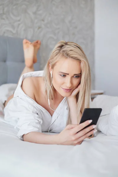 Szép, mosolygós szőke nő fehér ágyban fekve, és egy okostelefon segítségével — Stock Fotó