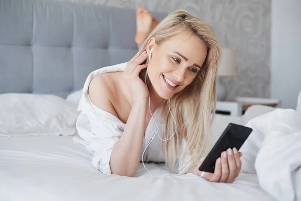 Mujer rubia hermosa y sonriente acostada en una cama blanca y usando un teléfono inteligente —  Fotos de Stock