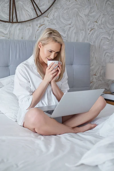 Gelukkig mooie vrouw die werkt op een laptop zitten op het bed in het huis. — Stockfoto