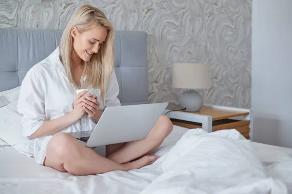 Mulher bonita feliz trabalhando em um laptop sentado na cama na casa . — Fotografia de Stock