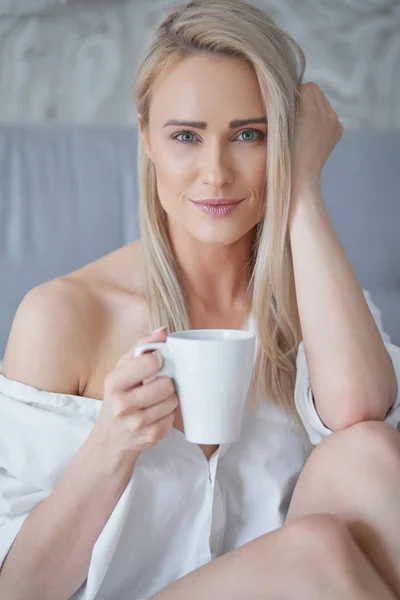 Mooie blonde vrouw drinken van een kopje koffie zittend op haar bed thuis — Stockfoto