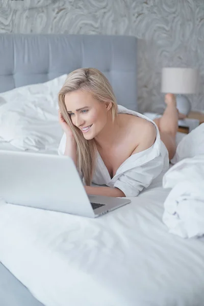 Een glimlachende blonde vrouw liggend op het bed voor haar laptop — Stockfoto