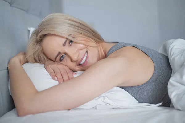 Χαμογελώντας casual ξανθιά ξαπλωμένη στο κρεβάτι της σε φωτεινό υπνοδωμάτιο — Φωτογραφία Αρχείου