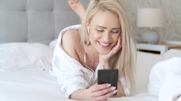 아름 다운, 웃는 금발 여자 흰 침대에 누워 및 smartphone을 사용 하 여 — 비디오