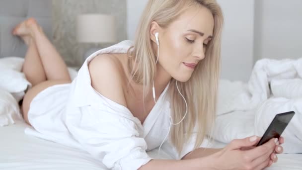 Schöne, lächelnde blonde Frau, die im weißen Bett liegt und ein Smartphone benutzt — Stockvideo